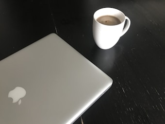 A closed silver laptop with a prominent logo on its lid is positioned on a dark surface. Next to the laptop is a white textured mug filled with a hot beverage, likely coffee, creating a simple and minimalist workspace setting.