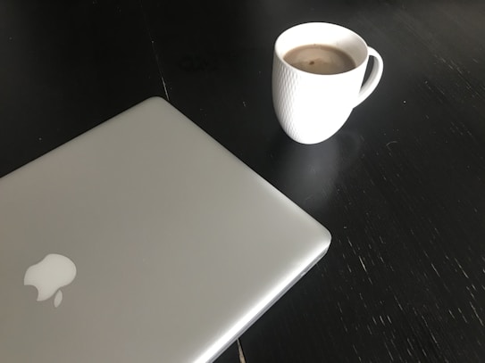 A closed silver laptop with a prominent logo on its lid is positioned on a dark surface. Next to the laptop is a white textured mug filled with a hot beverage, likely coffee, creating a simple and minimalist workspace setting.
