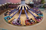 A vibrant amusement park ride featuring colorful vehicles arranged in a circular pattern around a central spinning mechanism. The ride is adorned with purple and gold accents, and each vehicle has unique decorations and seating for two passengers. The area is surrounded by decorative fencing and an artistically painted backdrop.
