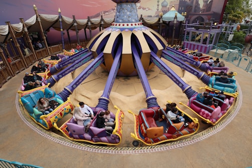 A vibrant amusement park ride featuring colorful vehicles arranged in a circular pattern around a central spinning mechanism. The ride is adorned with purple and gold accents, and each vehicle has unique decorations and seating for two passengers. The area is surrounded by decorative fencing and an artistically painted backdrop.