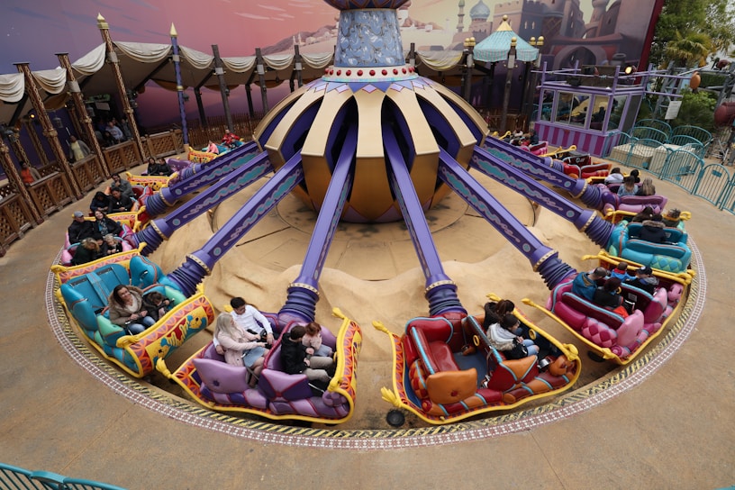 A vibrant amusement park ride featuring colorful vehicles arranged in a circular pattern around a central spinning mechanism. The ride is adorned with purple and gold accents, and each vehicle has unique decorations and seating for two passengers. The area is surrounded by decorative fencing and an artistically painted backdrop.