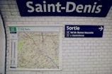 A white tiled wall in a subway station displays a map of Paris alongside signs with directions. The word 'Saint-Denis' is prominently featured at the top. To the right, a blue exit sign with the text 'Sortie' points towards directions with numbers 4 and 5.