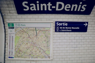 A white tiled wall in a subway station displays a map of Paris alongside signs with directions. The word 'Saint-Denis' is prominently featured at the top. To the right, a blue exit sign with the text 'Sortie' points towards directions with numbers 4 and 5.