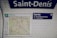 A white tiled wall in a subway station displays a map of Paris alongside signs with directions. The word 'Saint-Denis' is prominently featured at the top. To the right, a blue exit sign with the text 'Sortie' points towards directions with numbers 4 and 5.