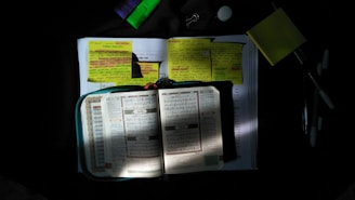 Books and notebooks are open on a surface, illuminated by a focused light. Yellow sticky notes with handwritten text are placed on the pages, and writing tools such as pens and highlighters are nearby. The scene is set against a dark background, creating a contrast with the lit area.