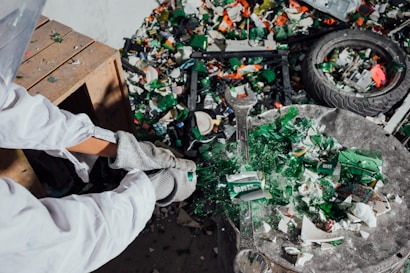 A person wearing protective gloves and a white suit is working with broken pieces of green glass, likely recycling or sorting materials. A large wrench is visible among the debris. A wooden table or shelf is present on the left side, and a tire and various pieces of metal and plastic are scattered around.