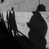 Shadows of people are cast on a textured wall, with each individual having a distinct silhouette, possibly indicating a variety of postures and actions. The play of light and shadow creates an intriguing visual narrative.