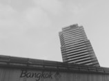 A towering rectangular skyscraper with a modern design, featuring alternating dark and light horizontal stripes, ascends against a pale sky. In the foreground, a structure displays the text 'Bangkok, City of Life,' indicating an urban setting.