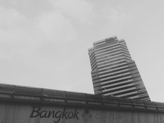 A towering rectangular skyscraper with a modern design, featuring alternating dark and light horizontal stripes, ascends against a pale sky. In the foreground, a structure displays the text 'Bangkok, City of Life,' indicating an urban setting.
