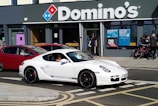 A white sports car with black rims and the logo of a popular car brand is parked on the street in front of a Domino's pizza outlet. The store has large glass windows with advertisement posters and a few people are standing near the entrance. A red car is behind the white car, and some people with bicycles are nearby.