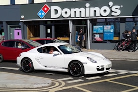 A white sports car with black rims and the logo of a popular car brand is parked on the street in front of a Domino's pizza outlet. The store has large glass windows with advertisement posters and a few people are standing near the entrance. A red car is behind the white car, and some people with bicycles are nearby.