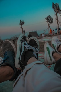 Feet wearing casual shoes relax on a patterned mat. In the background, there are structures resembling trees with abstract top parts. The sky is a clear blue, and there are people gathered at a distance, creating a laid-back atmosphere.
