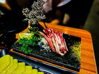 A black rectangular plate holds slices of raw meat, garnished with chopped parsley, a slice of lime, and decorative flowers. Next to it, another plate contains neatly arranged slices of yellow pickled vegetables. The surface is wooden, creating a contrasting backdrop for the vibrant colors of the dish.