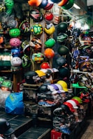 A vibrant display of various helmets in a shop, showcasing an array of colors and designs arranged neatly on shelves and hanging on walls. Both full-face and open helmets are visible, along with an assortment of patterns and styles. The shop is well-lit, with a bustling, busy feel.