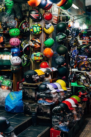 A vibrant display of various helmets in a shop, showcasing an array of colors and designs arranged neatly on shelves and hanging on walls. Both full-face and open helmets are visible, along with an assortment of patterns and styles. The shop is well-lit, with a bustling, busy feel.
