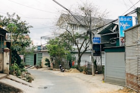 A narrow, paved street in a residential area lined with houses and shops with metal shutters. Overhead electrical wires are visible, and the area is surrounded by trees and greenery. There are several signs in Vietnamese along the street.