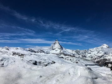 A majestic snow-covered mountain range with a prominent peak surrounded by a clear blue sky and scattered clouds. The landscape is covered with pristine white snow, creating a serene and expansive alpine vista.
