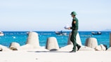 A person in a green uniform and helmet is walking along a coastal area holding papers. In the background, there are large concrete breakwaters and several small boats floating on a bright blue sea with distant hills visible under a clear sky.