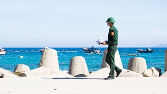 A person in a green uniform and helmet is walking along a coastal area holding papers. In the background, there are large concrete breakwaters and several small boats floating on a bright blue sea with distant hills visible under a clear sky.