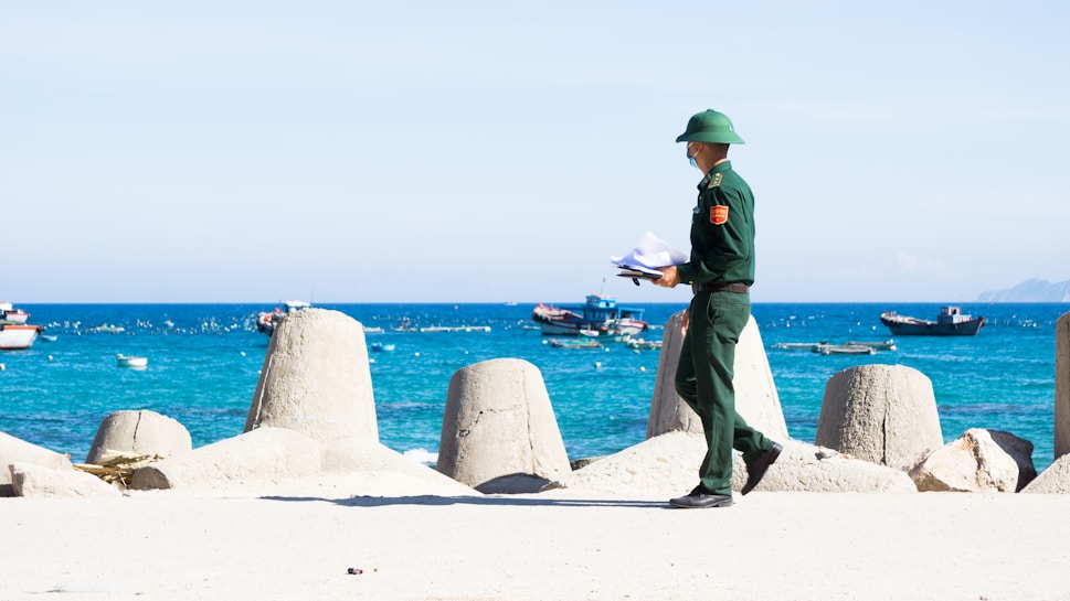 A person in a green uniform and helmet is walking along a coastal area holding papers. In the background, there are large concrete breakwaters and several small boats floating on a bright blue sea with distant hills visible under a clear sky.