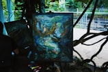 An abstract painting on an easel with swirling colors and patterns, possibly depicting faces and figures interwoven in a chaotic manner. The painting is set outdoors with trees and a wrought iron fence in the background. A person is seated near the painting with art supplies scattered around them.