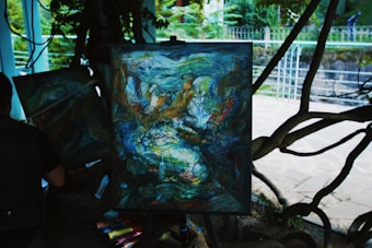 An abstract painting on an easel with swirling colors and patterns, possibly depicting faces and figures interwoven in a chaotic manner. The painting is set outdoors with trees and a wrought iron fence in the background. A person is seated near the painting with art supplies scattered around them.