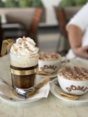 A close-up of two coffee beverages served in a cafe setting. In the foreground, there is an elegant glass cup with metal accents, filled with a coffee topped with whipped cream and cocoa powder. Next to it, a cappuccino cup with a frothy top covered in cocoa, branded with the name 'DOME', rests on a saucer accompanied by a cookie. Both cups are placed on white plates with spoons.