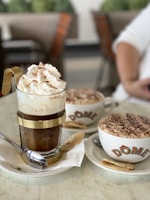 A close-up of two coffee beverages served in a cafe setting. In the foreground, there is an elegant glass cup with metal accents, filled with a coffee topped with whipped cream and cocoa powder. Next to it, a cappuccino cup with a frothy top covered in cocoa, branded with the name 'DOME', rests on a saucer accompanied by a cookie. Both cups are placed on white plates with spoons.