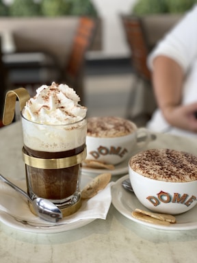 A close-up of two coffee beverages served in a cafe setting. In the foreground, there is an elegant glass cup with metal accents, filled with a coffee topped with whipped cream and cocoa powder. Next to it, a cappuccino cup with a frothy top covered in cocoa, branded with the name 'DOME', rests on a saucer accompanied by a cookie. Both cups are placed on white plates with spoons.
