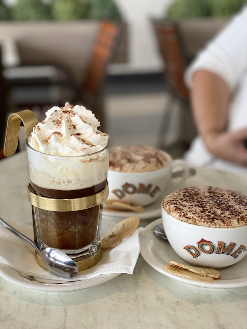 A close-up of two coffee beverages served in a cafe setting. In the foreground, there is an elegant glass cup with metal accents, filled with a coffee topped with whipped cream and cocoa powder. Next to it, a cappuccino cup with a frothy top covered in cocoa, branded with the name 'DOME', rests on a saucer accompanied by a cookie. Both cups are placed on white plates with spoons.