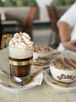 A close-up of two coffee beverages served in a cafe setting. In the foreground, there is an elegant glass cup with metal accents, filled with a coffee topped with whipped cream and cocoa powder. Next to it, a cappuccino cup with a frothy top covered in cocoa, branded with the name 'DOME', rests on a saucer accompanied by a cookie. Both cups are placed on white plates with spoons.