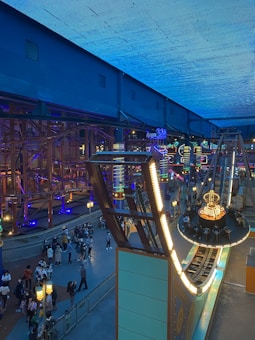 An indoor amusement park setting featuring a large, illuminated swing ride with people seated, surrounded by vibrant neon lights. The scene includes a crowd of people walking and observing the attractions, with the ceiling covered in a bright blue hue.
