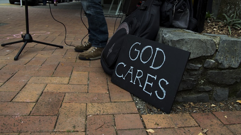 A sidewalk features a handwritten sign saying 'God Cares' leaning against a stone wall. Part of a person's legs in jeans and brown shoes are visible beside the sign. A microphone stand and wires are present, indicating a possible street performance setup. A black backpack is also placed near the wall.