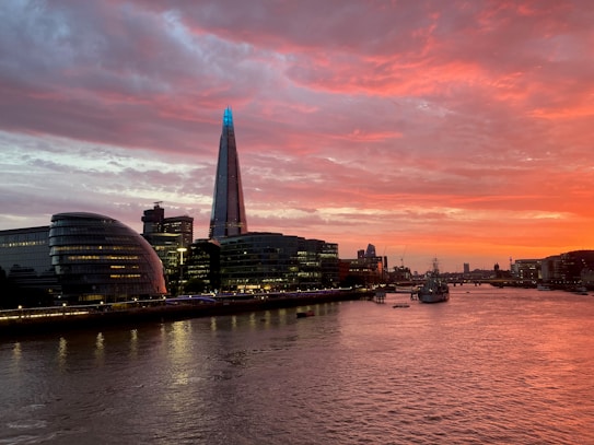 A stunning cityscape at dusk with a prominent skyscraper illuminated against a vibrant sunset. The sky is filled with shades of orange, red, and pink, creating a picturesque backdrop over a river lined with modern buildings. Streetlights add a warm glow along the riverbank.