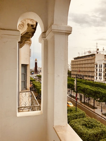 An elegant balcony with ornate wrought iron railings overlooks a bustling city street lined with green trees. In the distance, a tall tower stands against a cloudy sky, surrounded by various buildings with antennas atop.