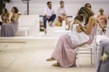 A woman in a pastel pink dress holds a young child in a white dress on her lap as they sit on a white chair. The setting appears to be an outdoor event with several people sitting and chatting in the background, creating a relaxed and intimate atmosphere. There are petals scattered on the floor, suggesting a celebratory occasion.
