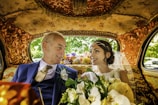 An elegantly dressed couple is sitting inside a lavishly decorated car. The interior features intricate gold patterns on the ceiling and seats. The woman, wearing a white wedding dress and veil, holds a bouquet of flowers. Both individuals are smiling warmly at each other, conveying a sense of joy and connection.