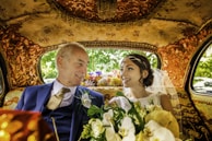 An elegantly dressed couple is sitting inside a lavishly decorated car. The interior features intricate gold patterns on the ceiling and seats. The woman, wearing a white wedding dress and veil, holds a bouquet of flowers. Both individuals are smiling warmly at each other, conveying a sense of joy and connection.