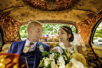 An elegantly dressed couple is sitting inside a lavishly decorated car. The interior features intricate gold patterns on the ceiling and seats. The woman, wearing a white wedding dress and veil, holds a bouquet of flowers. Both individuals are smiling warmly at each other, conveying a sense of joy and connection.