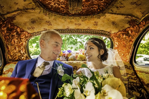 An elegantly dressed couple is sitting inside a lavishly decorated car. The interior features intricate gold patterns on the ceiling and seats. The woman, wearing a white wedding dress and veil, holds a bouquet of flowers. Both individuals are smiling warmly at each other, conveying a sense of joy and connection.