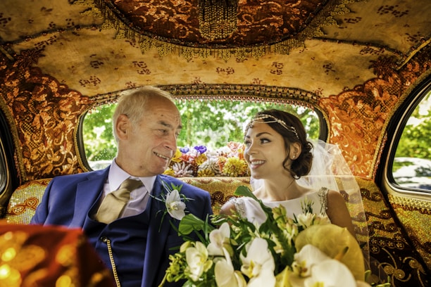 An elegantly dressed couple is sitting inside a lavishly decorated car. The interior features intricate gold patterns on the ceiling and seats. The woman, wearing a white wedding dress and veil, holds a bouquet of flowers. Both individuals are smiling warmly at each other, conveying a sense of joy and connection.