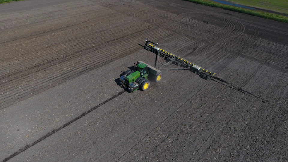Large rectangular field with rows of dirt patches, dominated by a green tractor plowing