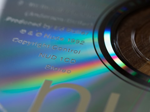 Close-up view of a compact disc with visible rainbow-like reflections and text printed on its surface. The text includes details such as 'Produced by Ed Buller', 'Nude 1992', 'Copyright Control', and 'Stereo'. The disc itself has a shiny, reflective surface with iridescent hues visible around the edges.