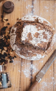 A rustic loaf of bread with a cracked, flour-dusted crust sits on a wooden surface. Surrounding the bread are scattered nuts and dried berries, with a small glass spice jar and a serrated knife nearby. Lightly sprinkled flour enhances the natural, homemade aesthetic.