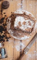 A rustic loaf of bread with a cracked, flour-dusted crust sits on a wooden surface. Surrounding the bread are scattered nuts and dried berries, with a small glass spice jar and a serrated knife nearby. Lightly sprinkled flour enhances the natural, homemade aesthetic.
