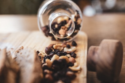 A glass jar is tipped over on its side, spilling out a mix of nuts and dried fruits onto a wooden cutting board. The assortment includes cashews, almonds, raisins, and hazelnuts, adding a rustic and natural vibe to the scene.