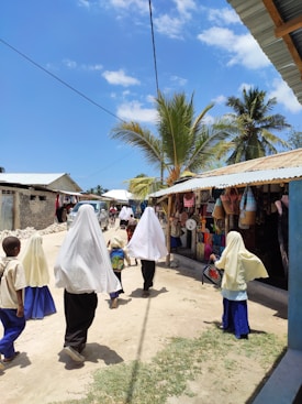 A group of children walks down a dirt street in a sunny village scene. The children are dressed in similar clothing, with head coverings and long skirts or pants. The street is bordered by small shops displaying various items, including hats and woven baskets. Palm trees and a clear blue sky dominate the background, creating a bright and vibrant atmosphere.