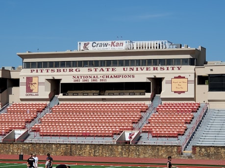 A stadium with tiers of empty red and gray seats is positioned in front of a tan building with multiple windows. The building is marked with the name 'Pittsburg State University' and indicates several national championship years. A large banner on the roof advertises 'Craw-Kan' along with services like fiber-optic internet and video. The university's gorilla mascot logo is visible on a pillar. People are walking on the athletic track in the foreground.