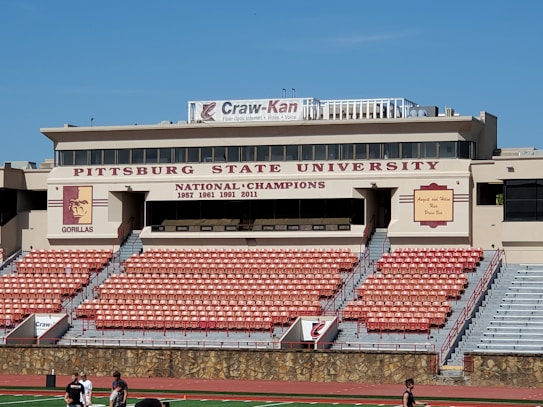 A stadium with tiers of empty red and gray seats is positioned in front of a tan building with multiple windows. The building is marked with the name 'Pittsburg State University' and indicates several national championship years. A large banner on the roof advertises 'Craw-Kan' along with services like fiber-optic internet and video. The university's gorilla mascot logo is visible on a pillar. People are walking on the athletic track in the foreground.