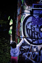 A child with outstretched arms reaches toward a large, graffiti-covered wall. The artwork on the wall features a mix of colors and abstract shapes, including shades of blue, green, and red. The setting appears to be outdoors, and the area around the child is dimly lit with natural light filtering through trees.